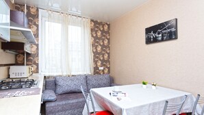 A compact kitchenette with a gas stove, a small refrigerator, and a coffee machine. There is a gray sofa with red chairs around a small dining table. A framed picture of a bridge is on the wall.
