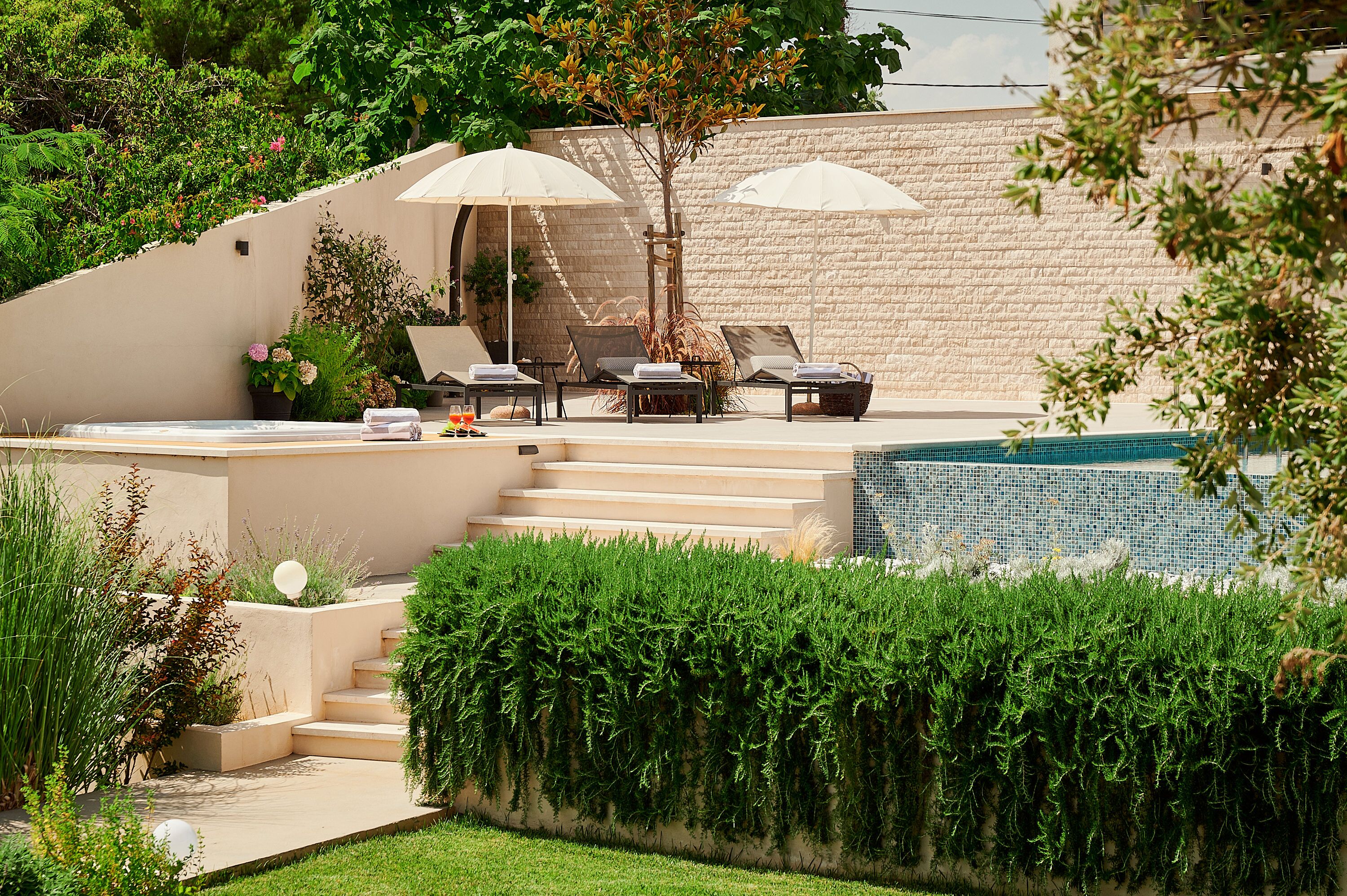 A pool area with lounge chairs and umbrellas, surrounded by greenery and a stone wall.