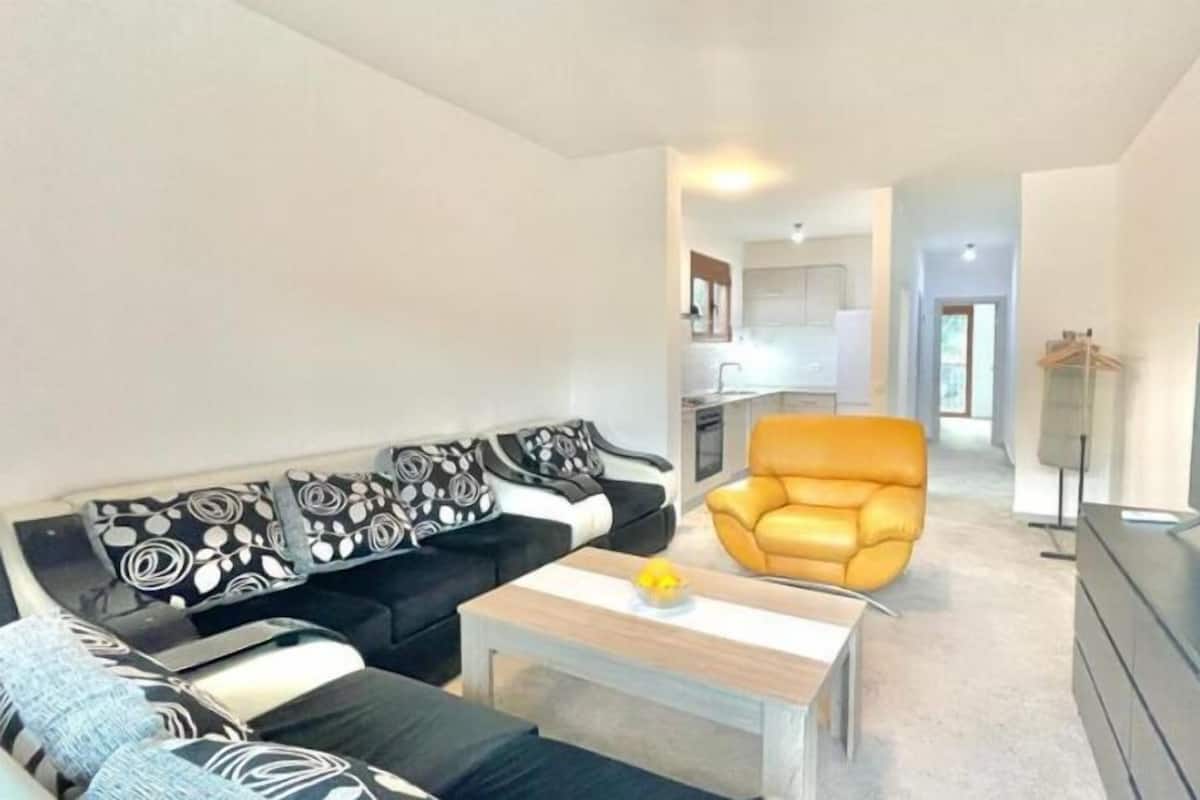 Una sala de estar moderna con un sofá de estampado blanco y negro, un sillón amarillo y una mesa de centro de madera. Un televisor de pantalla plana está montado en la pared.