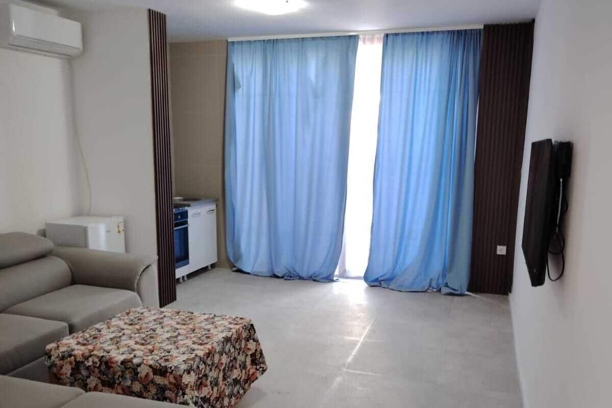 Una sala de estar con sofá, un taburete floral, un televisor de pantalla plana y una ventana con cortinas azules.