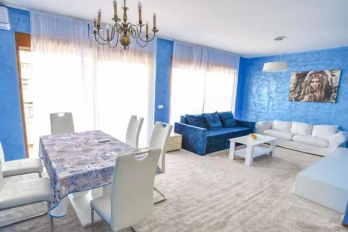 Un salón con pared azul, una zona de comedor con mantel estampado y una araña.
