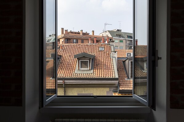 Vista a través de una ventana de un edificio con tejado de tejas, chimeneas visibles y una pequeña ventana con frontón triangular.