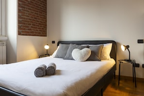 Una cama bien tendida con almohadas y toallas enrolladas, una pared de ladrillo y dos lámparas de noche.