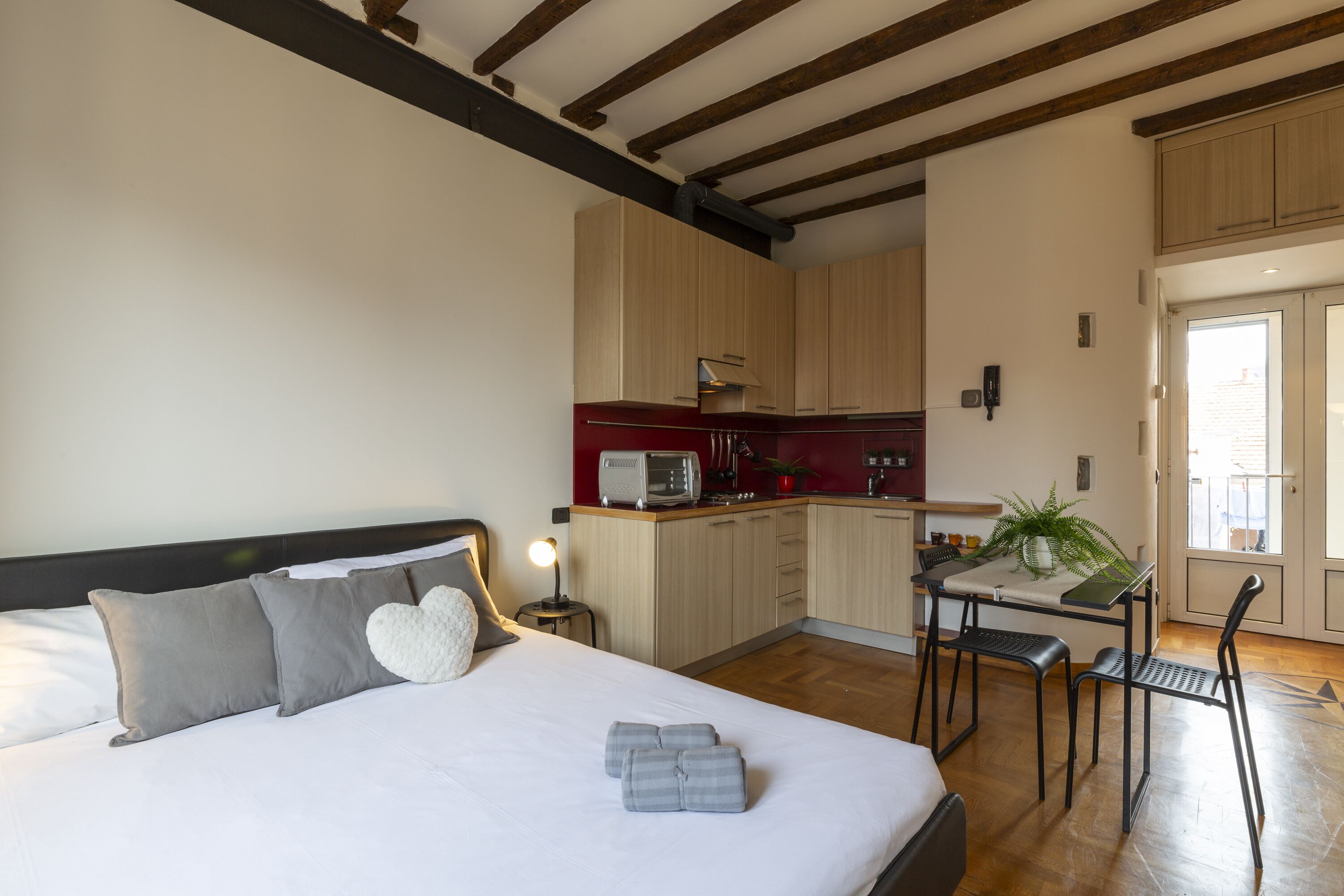 A bedroom with a bed, pillows, bedside table, and a night lamp. Adjacent is a kitchenette with a microwave, toaster, and cabinets. There is a dining table with chairs and a potted plant.