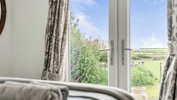 Une chambre avec une grande fenêtre, un lit avec une couverture enroulée, et une vue sur un paysage verdoyant.
