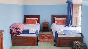 A room with two wooden beds, each with a blue canopy, white bedding, and orange pillows. There is a small nightstand with a lamp between the beds, and a patterned rug on the floor. - Riad Bounaylat (Addiss)