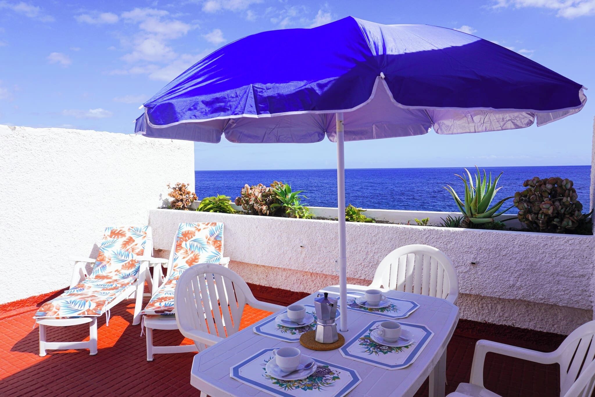 A terrace with a table set for two, white chairs, a blue umbrella, and a view of the sea.