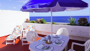 Terraço com mesa posta para dois, cadeiras brancas, um guarda-sol azul e vista para o mar.