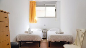 Quarto com duas camas, cómoda de madeira, uma cadeira, janela com cortinas e mesa de cabeceira.