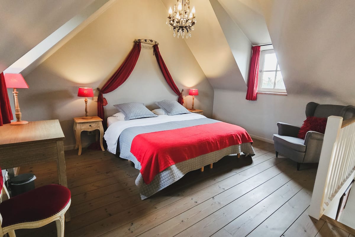 Une chambre avec un plafond en pente, un grand lit avec une literie rouge, deux tables de chevet avec des lampes, une chaise et une fenêtre avec des rideaux.