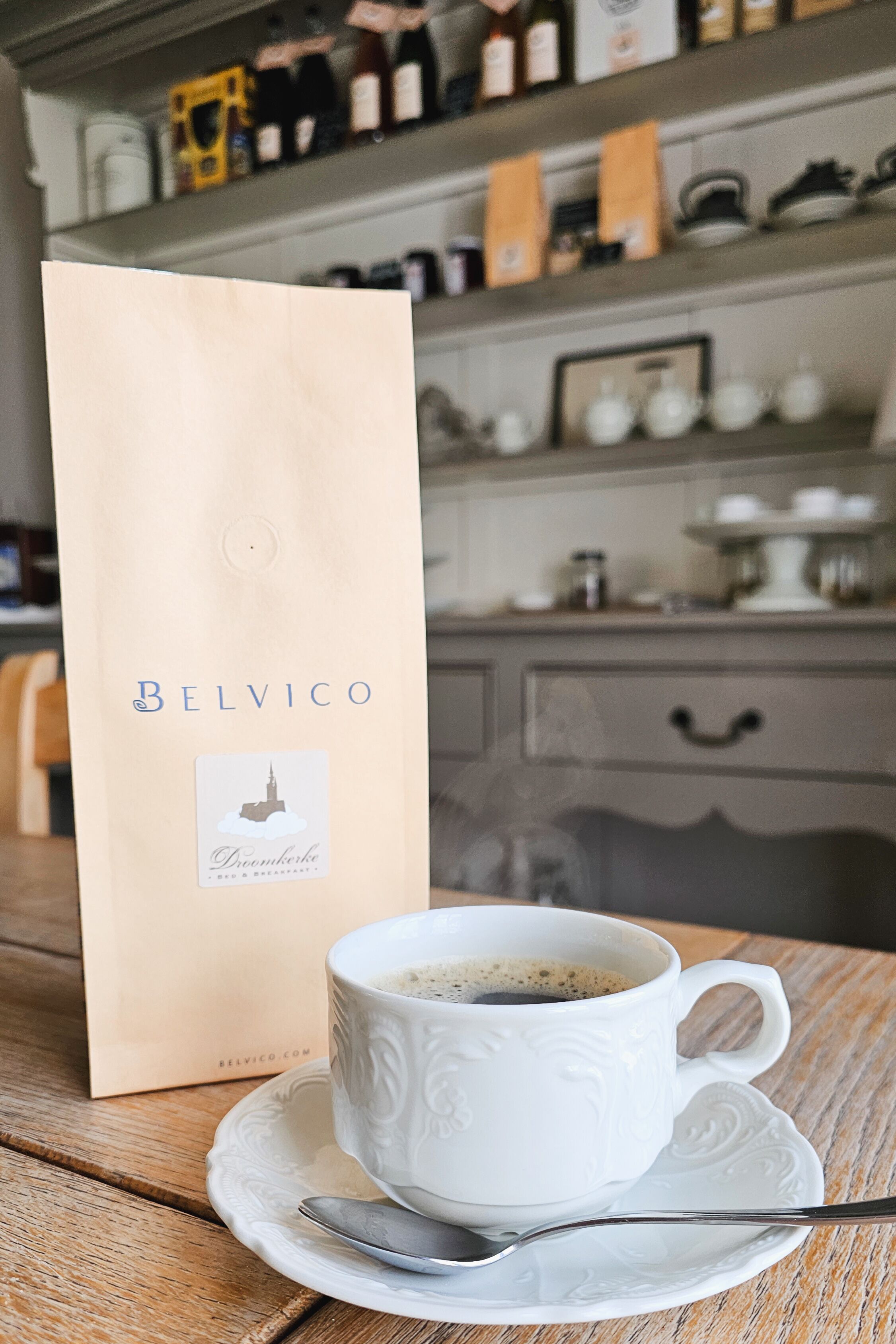 A white cup of coffee on a wooden table with a spoon, next to a Belvico branded paper bag.