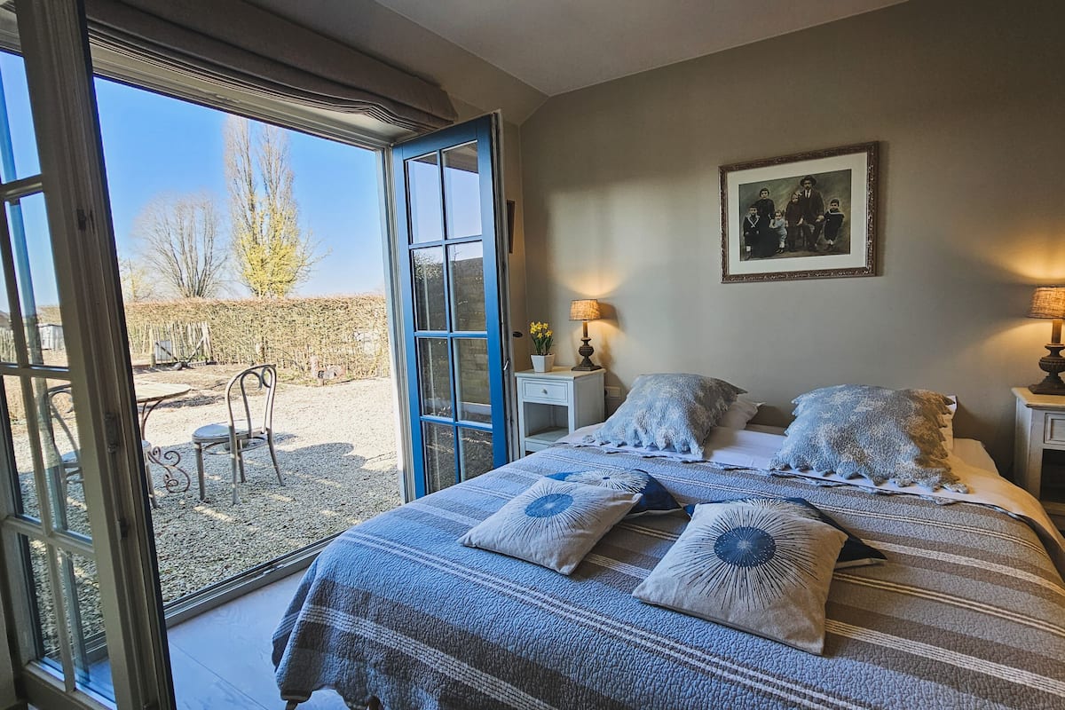 Une chambre avec un grand lit, deux tables de chevet, une commode, un portrait de famille encadré, et une vue sur un espace extérieur accessible par une porte ouverte, avec des chaises et un jardin.