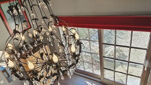 Ein geräumiges Zimmer mit einem großen Kronleuchter, einem Holztisch und Blick ins Freie durch große Fenster.