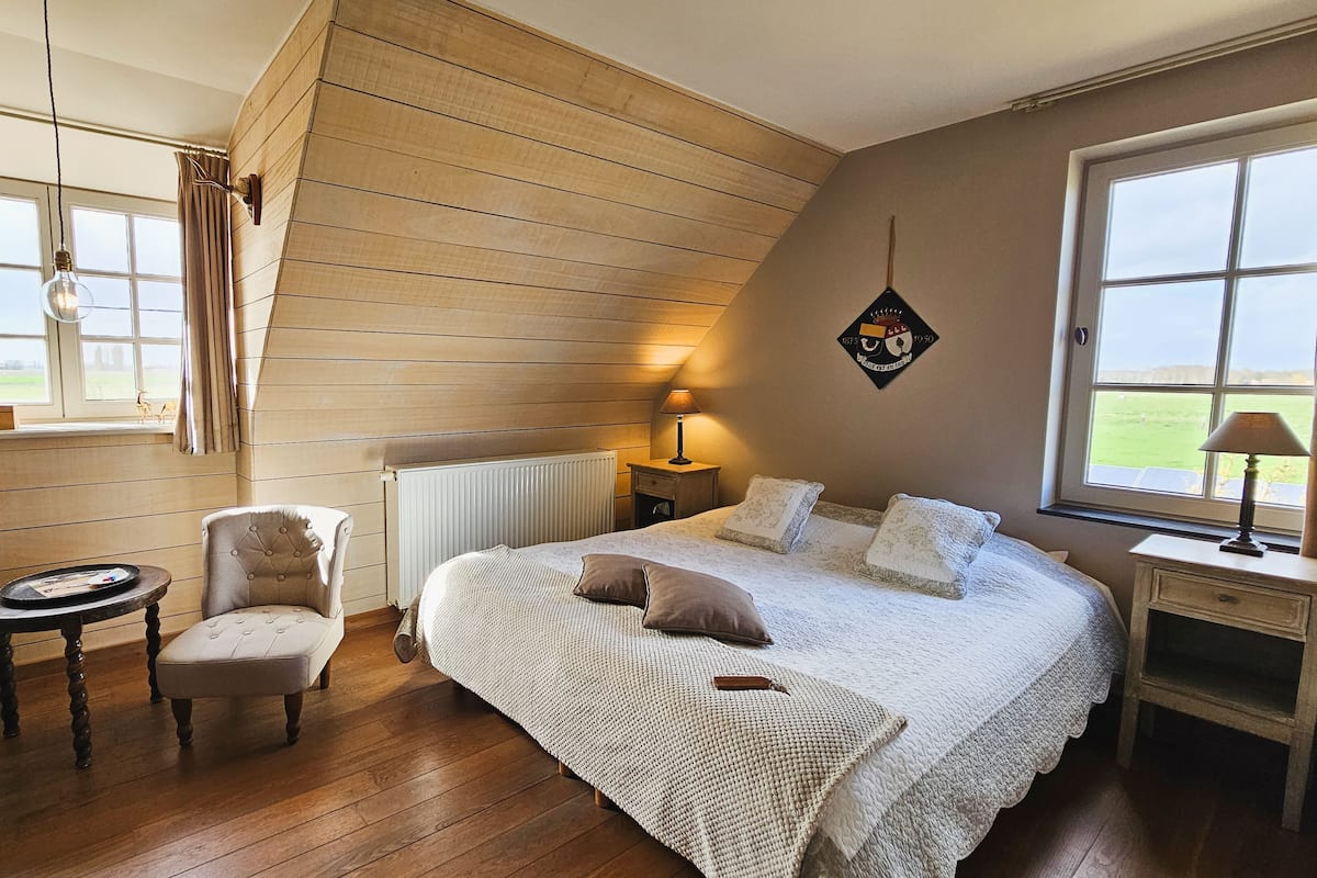 Une chambre confortable avec un plafond en pente en bois, un grand lit avec des oreillers, une chaise, une petite table et une fenêtre avec des rideaux.