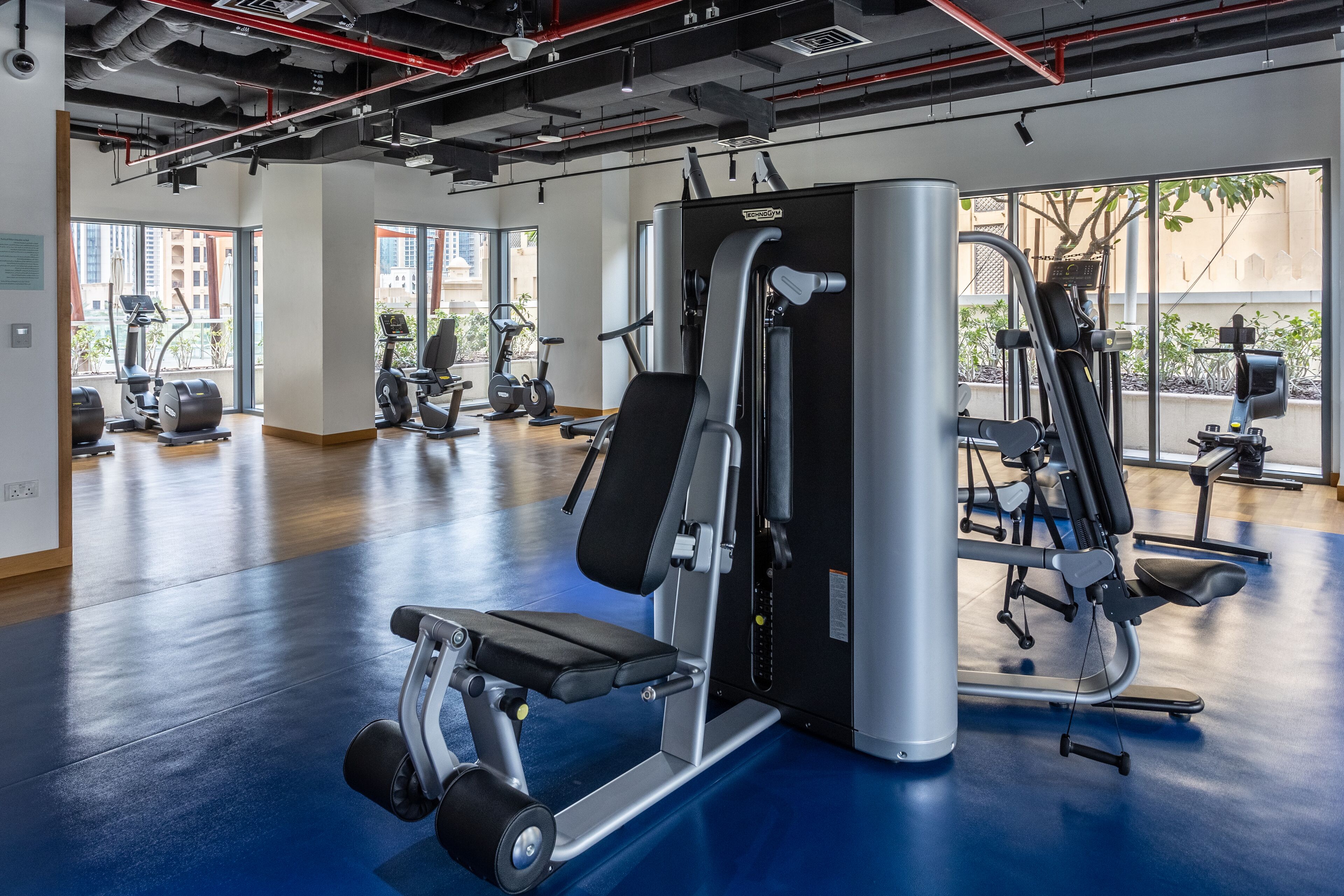 Une salle de sport équipée de divers appareils d’exercice, dotée de grandes fenêtres et d’un sol bleu.