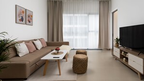 A modern living room with a sofa, coffee table, ottoman, and a TV on a wooden stand. - Waves - Chic Apartment With Dubai Skyline Views (Dubai)