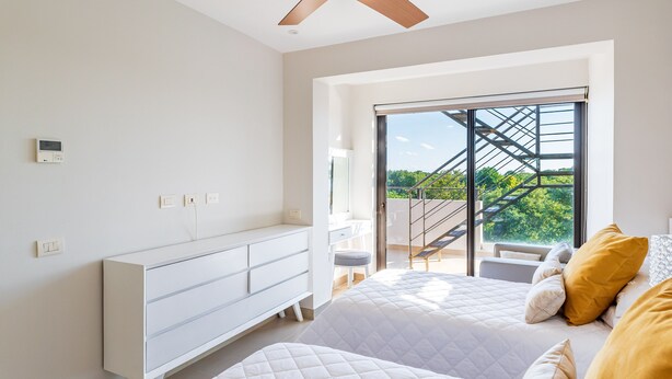 Quarto com cama, penteadeira, ventilador de teto, janela com vista para vegetação e varanda.