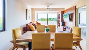 Uma sala de jantar moderna com uma mesa de madeira e cadeiras amarelas, um ventilador de teto e vista para o verde lá fora.