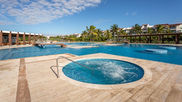 A large outdoor swimming pool with multiple sections, including a hot tub, surrounded by palm trees and a clear blue sky.