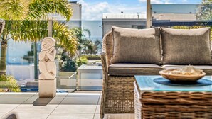 A wicker sofa with cushions and a small table with a bowl, set on a balcony with a view of palm trees and buildings. - 100 steps to the beach. Our modern Sunshine Coast home accommodates 12 guests (Alexandra Headland)