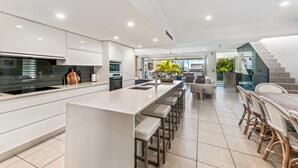 A modern kitchen with a central island, bar stools, and a dining area with wicker furniture. - 100 steps to the beach. Our modern Sunshine Coast home accommodates 12 guests (Alexandra Headland)