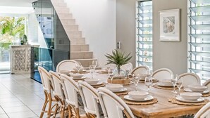 A dining area with a wooden table set for six, surrounded by wicker chairs. A staircase is visible in the background, leading to an upper level. - 100 steps to the beach. Our modern Sunshine Coast home accommodates 12 guests (Alexandra Headland)