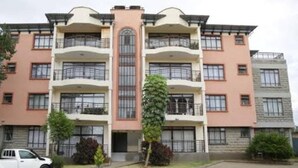 A multi-story residential building with balconies, a central entrance, and a parking area in front. - 3-bed Apartment in Nanyuki With Golf Course Views (Nanyuki)