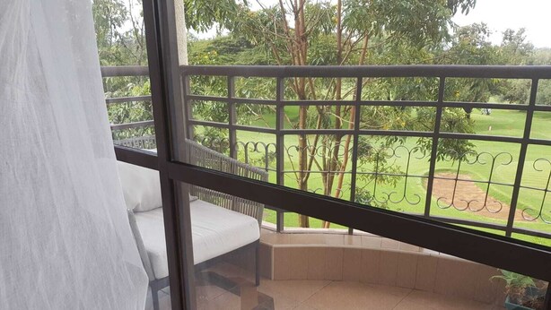 A balcony with a white cushioned seating area, a patterned tile floor, and a view of a green landscape with trees and a golf course. - 3-bed Apartment in Nanyuki With Golf Course Views (Nanyuki)