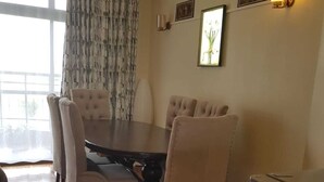 A dining area with a wooden herringbone floor, a dark wooden table, and four upholstered chairs. There is a window with patterned curtains, a wall-mounted light fixture, and a framed picture above the table. - 3-bed Apartment in Nanyuki With Golf Course Views (Nanyuki)