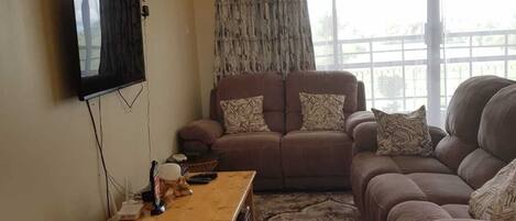 A living room with a sofa set, a television mounted on the wall, and a window with curtains.