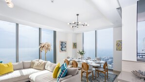 A modern living room with a large sectional sofa, dining area, and large windows offering a city view. - Waves - Sea View Luxury 3 Bedroom Apartment With Balcony (Dubai)