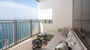 A balcony with a sofa, a small table, and a magazine. There is a view of the sea and buildings in the distance. - Waves - Sea View Luxury 3 Bedroom Apartment With Balcony (Dubai)