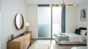 A modern bedroom with a large window, a wooden console table, a bed with white bedding, and a chandelier. - Waves - Sea View Luxury 3 Bedroom Apartment With Balcony (Dubai)