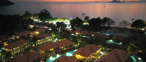 Un resort con varias bungalows sobre el agua, una piscina central y vistas al mar al atardecer.