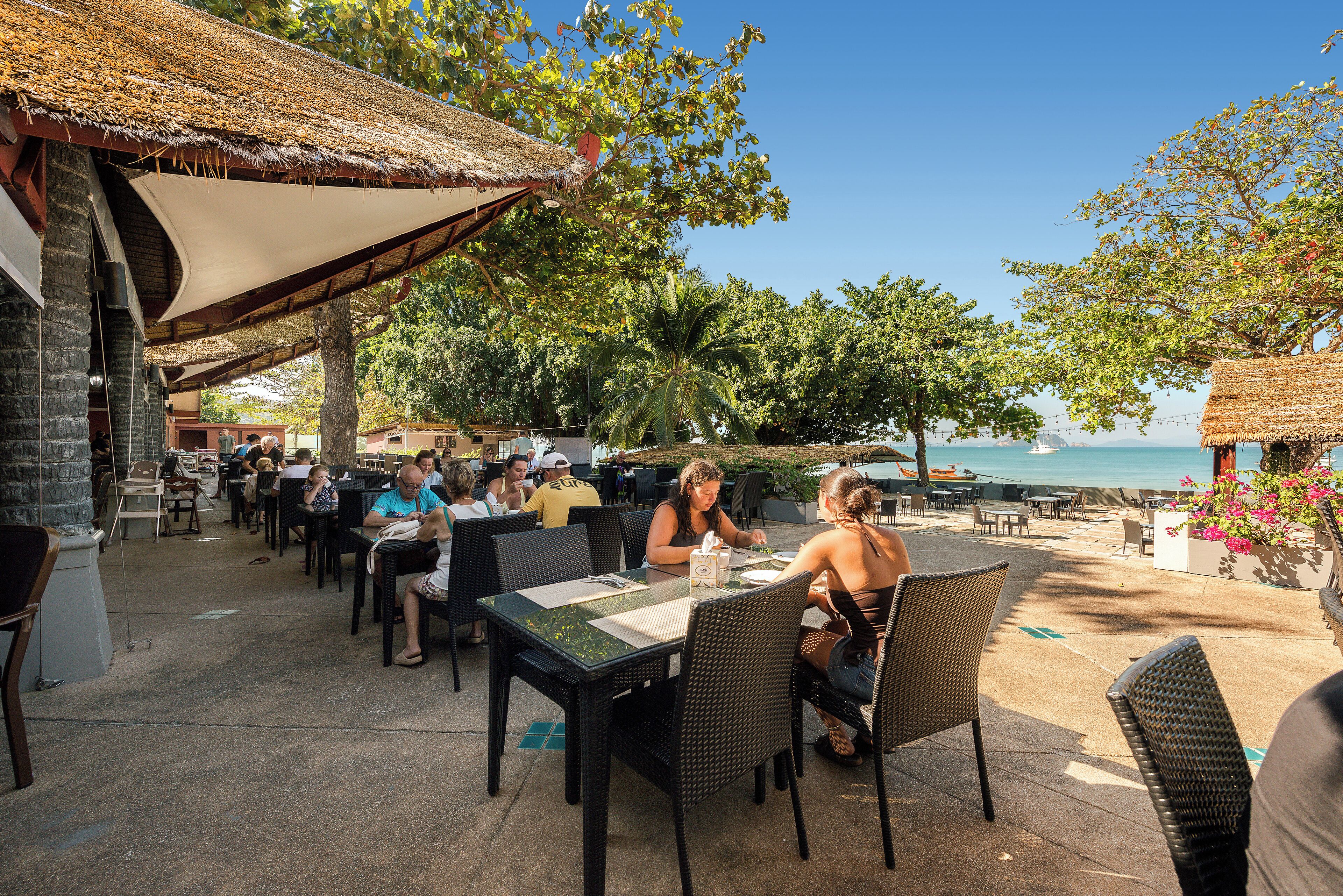 Un espace de restauration en plein air, avec des tables et des chaises, un toit de chaume et une vue sur la mer.