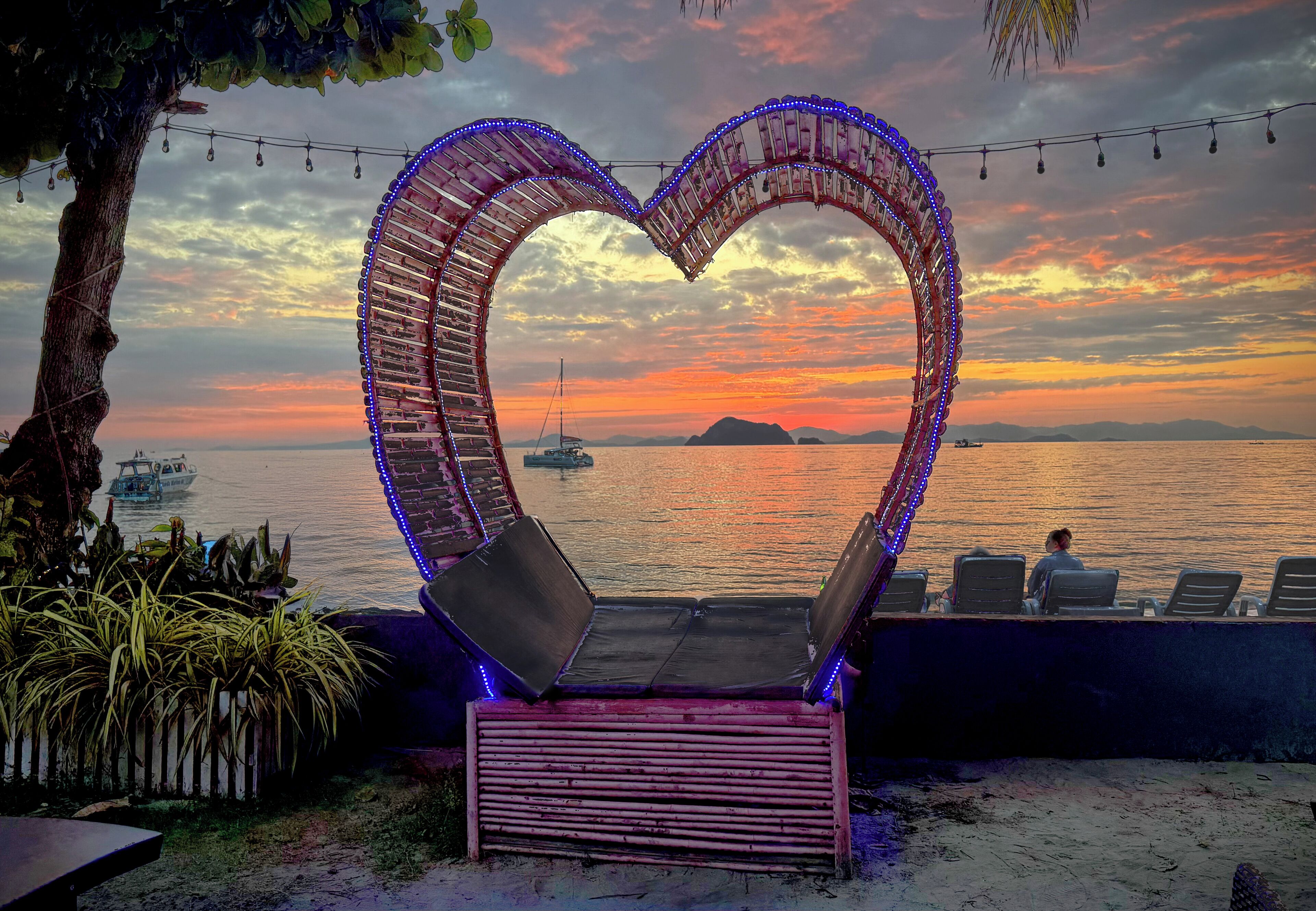 A heart-shaped structure with seating, overlooking a body of water with boats, at sunset.