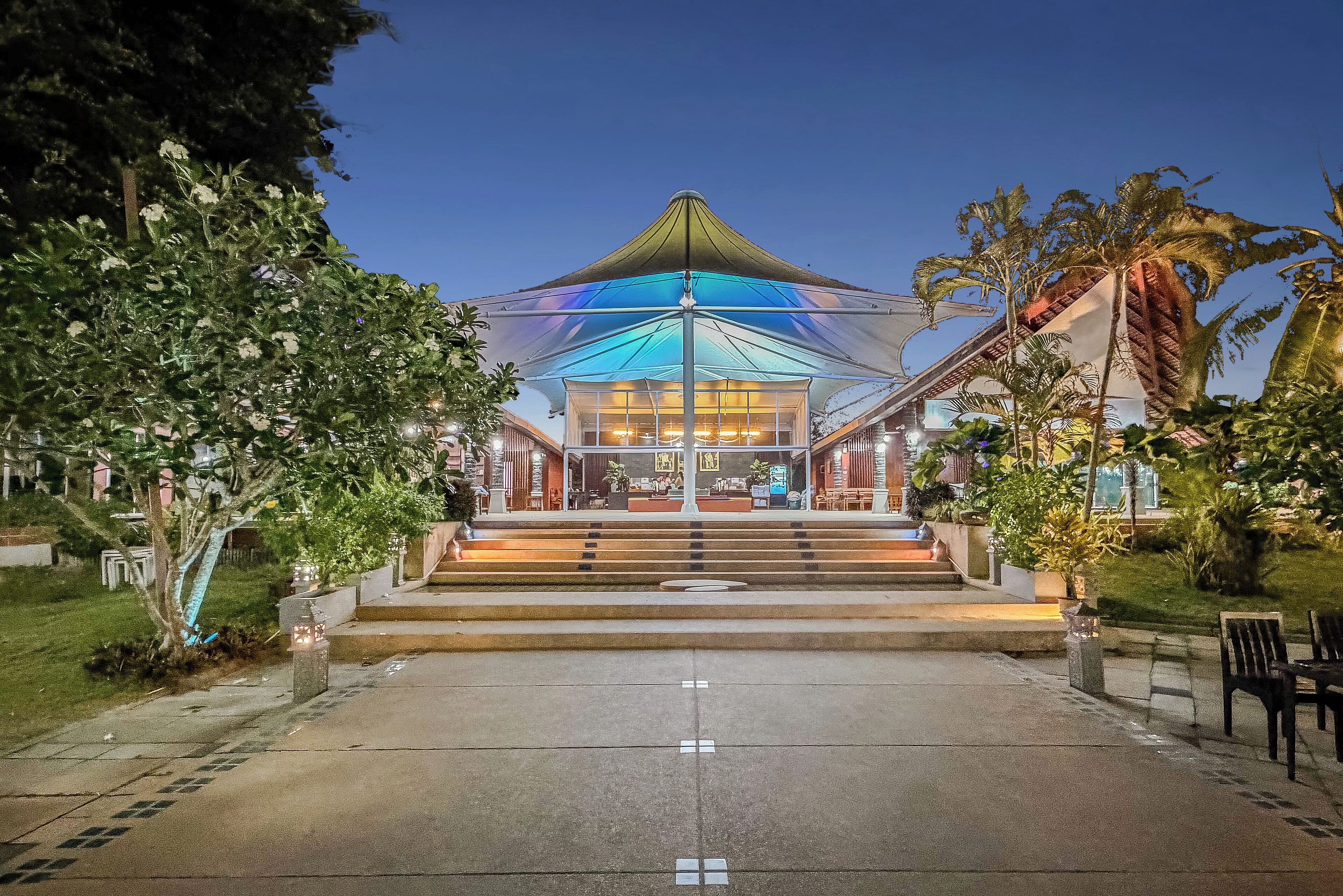 A building with a large, colorful canopy structure, a staircase leading to the entrance, and a well-maintained garden with palm trees.