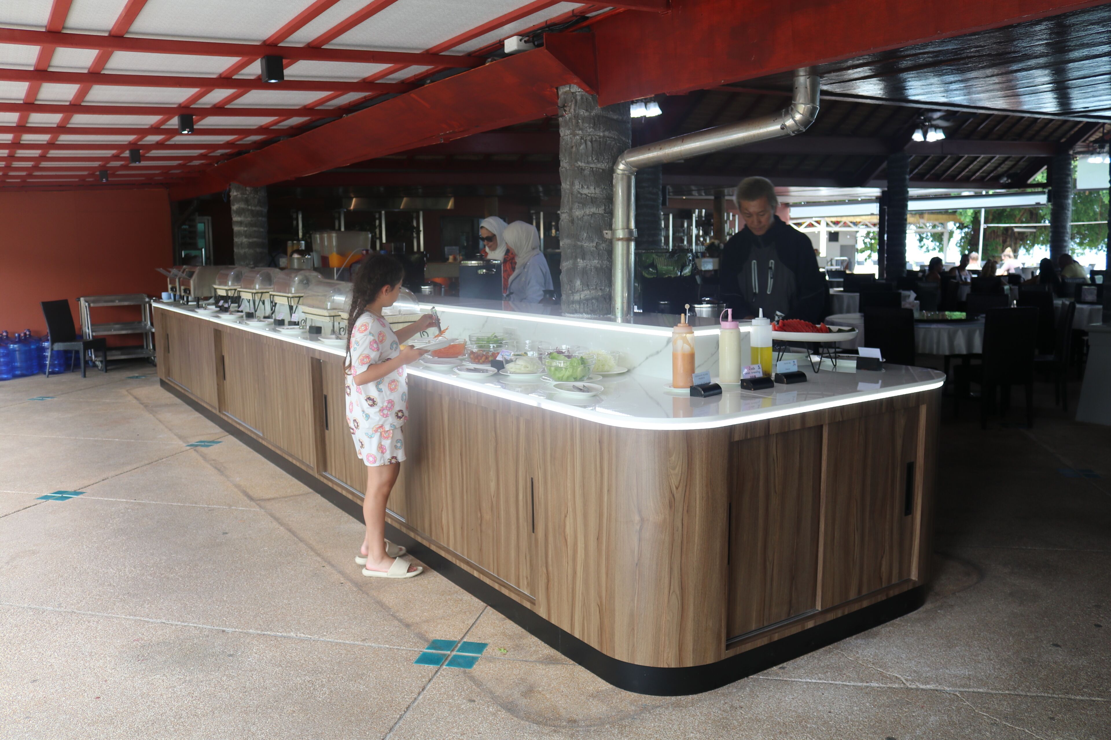 A buffet counter with a wooden finish, a person serving food, and a counter with various dishes and condiments.