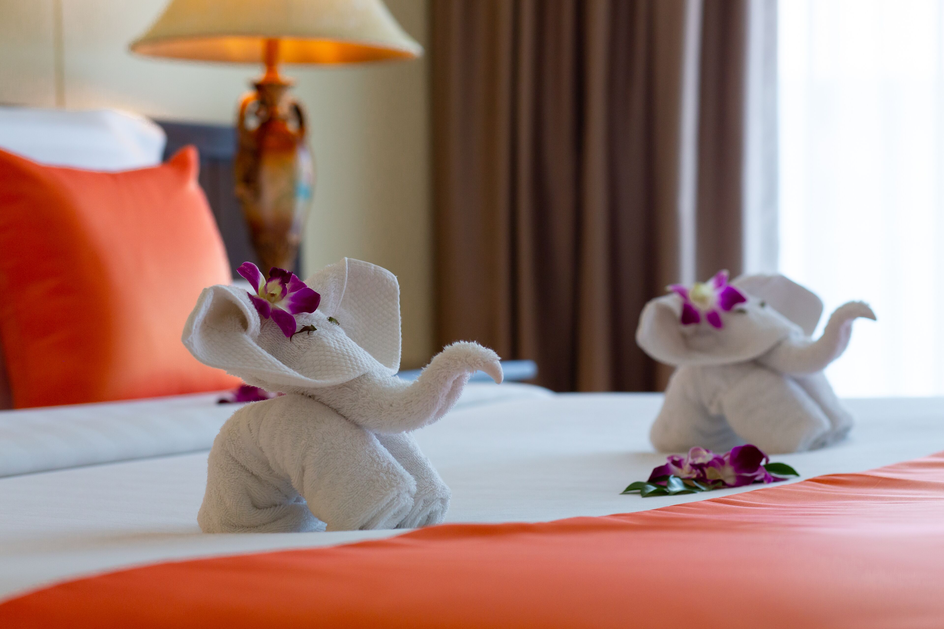 A hotel room with two plush elephant-shaped pillows on the bed, a lamp, and a curtain in the background.