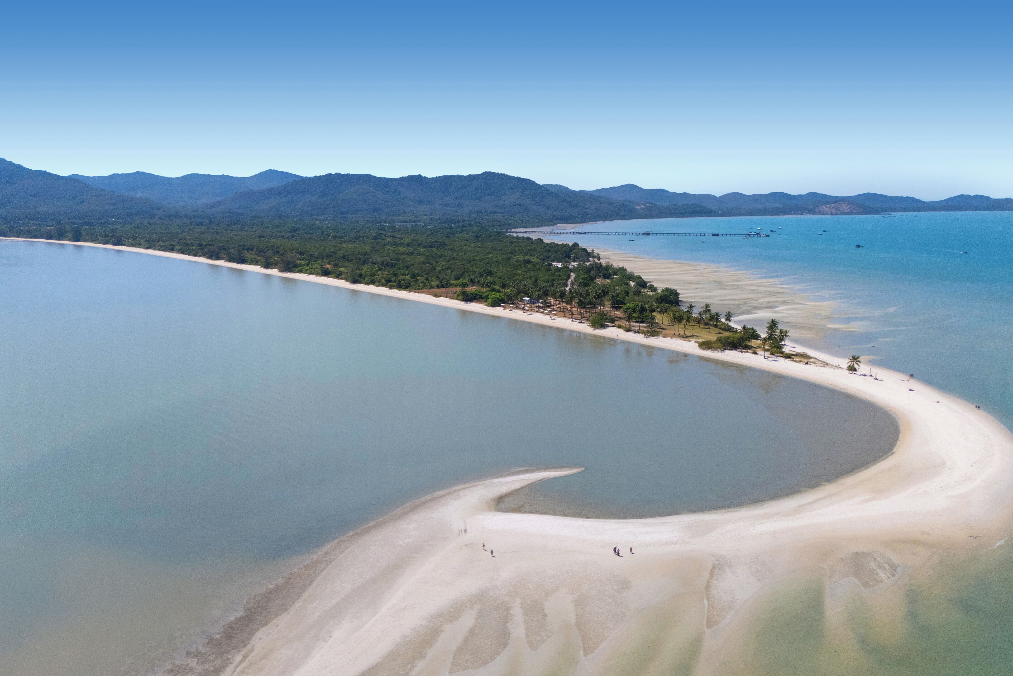 Vista aérea de una zona costera con playa arenosa, aguas azul turquesa y una península boscosa.