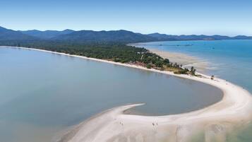 An aerial view of a coastal area with a sandy beach, clear blue waters, and a forested peninsula.