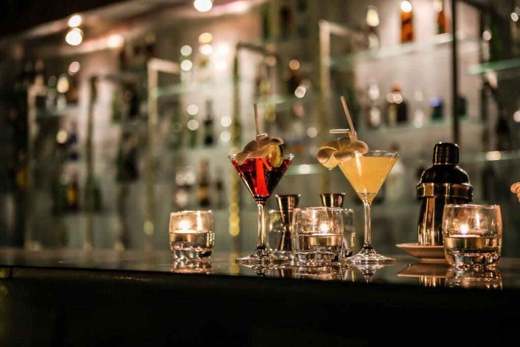 Eine Bar mit verschiedenen Gläsern und Flaschen, darunter ein Martini-Glas und ein Highball-Glas, mit unscharfem Hintergrund aus Regalen und Flaschen.