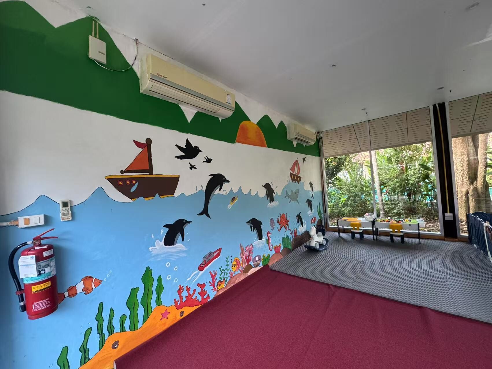 Une pièce décorée d’une fresque représentant la vie marine, équipée d’un extincteur et dotée d’une fenêtre donnant sur un paysage verdoyant.