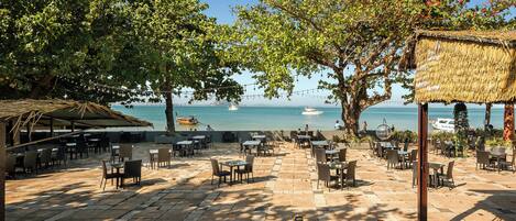An outdoor café with tables and chairs, overlooking a beach with boats and a clear sky.