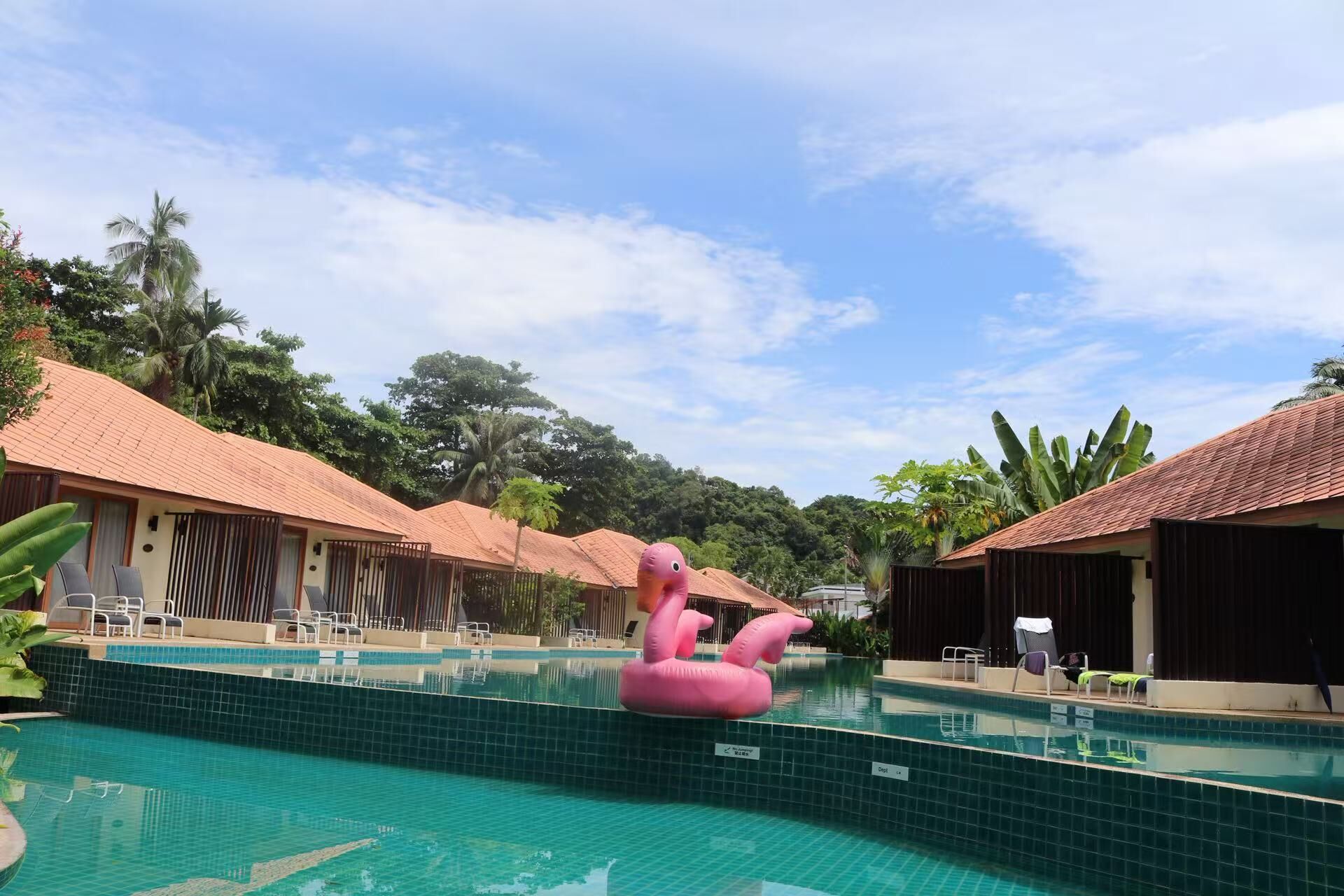 Ein Swimmingpool mit einem Flamingo-Schwimmpunkt, umgeben von Bungalows mit Ziegeldächern und tropischer Vegetation.