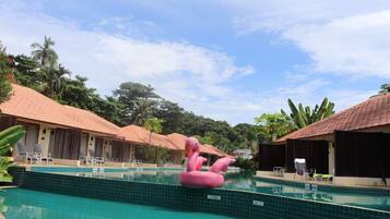 En swimmingpool med en lyserød flamingo flyder, omgivet af bungalows med flis tag og frodig, tropisk vegetation.