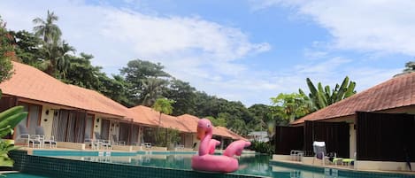 Una piscina con un flotador con forma de flamenco rosa, rodeada de edificios tipo cabaña con tejados de tejas y vegetación tropical.