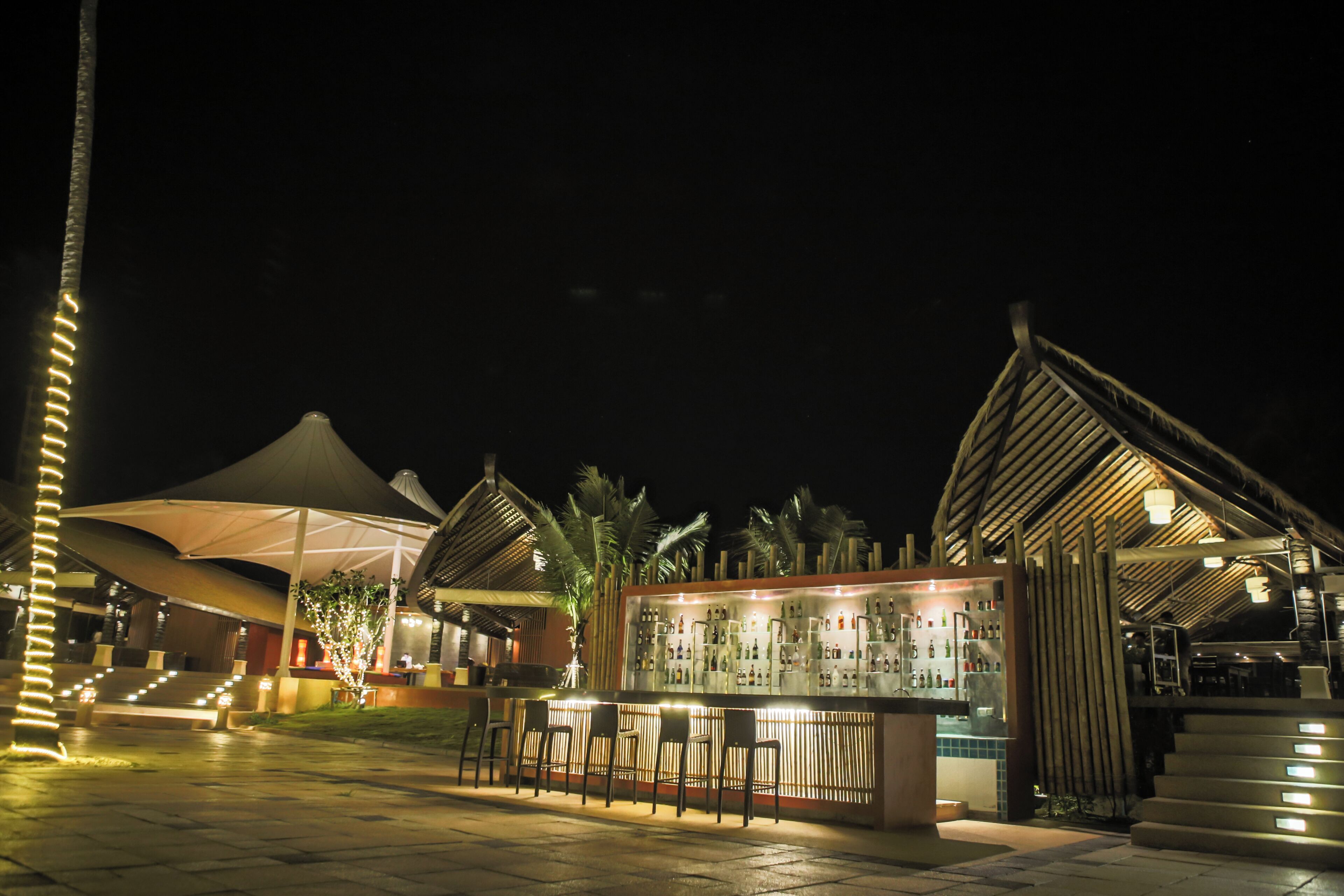 A bar area with seating and a counter, surrounded by palm trees and covered structures.