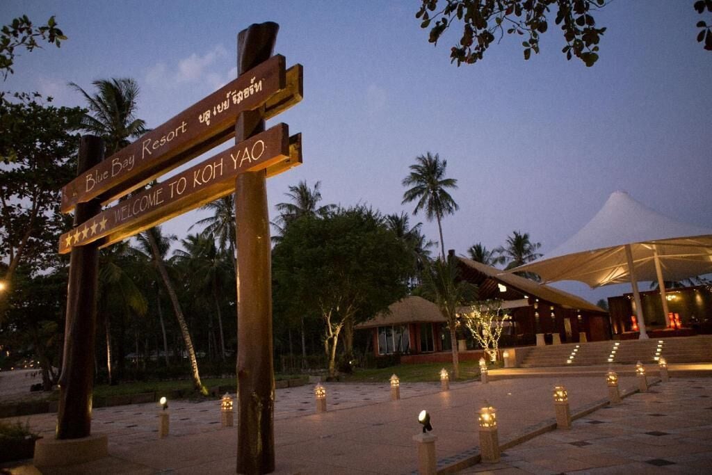 Ein hölzernes Wegweiser mit der Aufschrift 'Blue Bay Resort' und 'Willkommen auf Koh Yao', umgeben von Palmen und einem Gebäude mit einer Treppe.