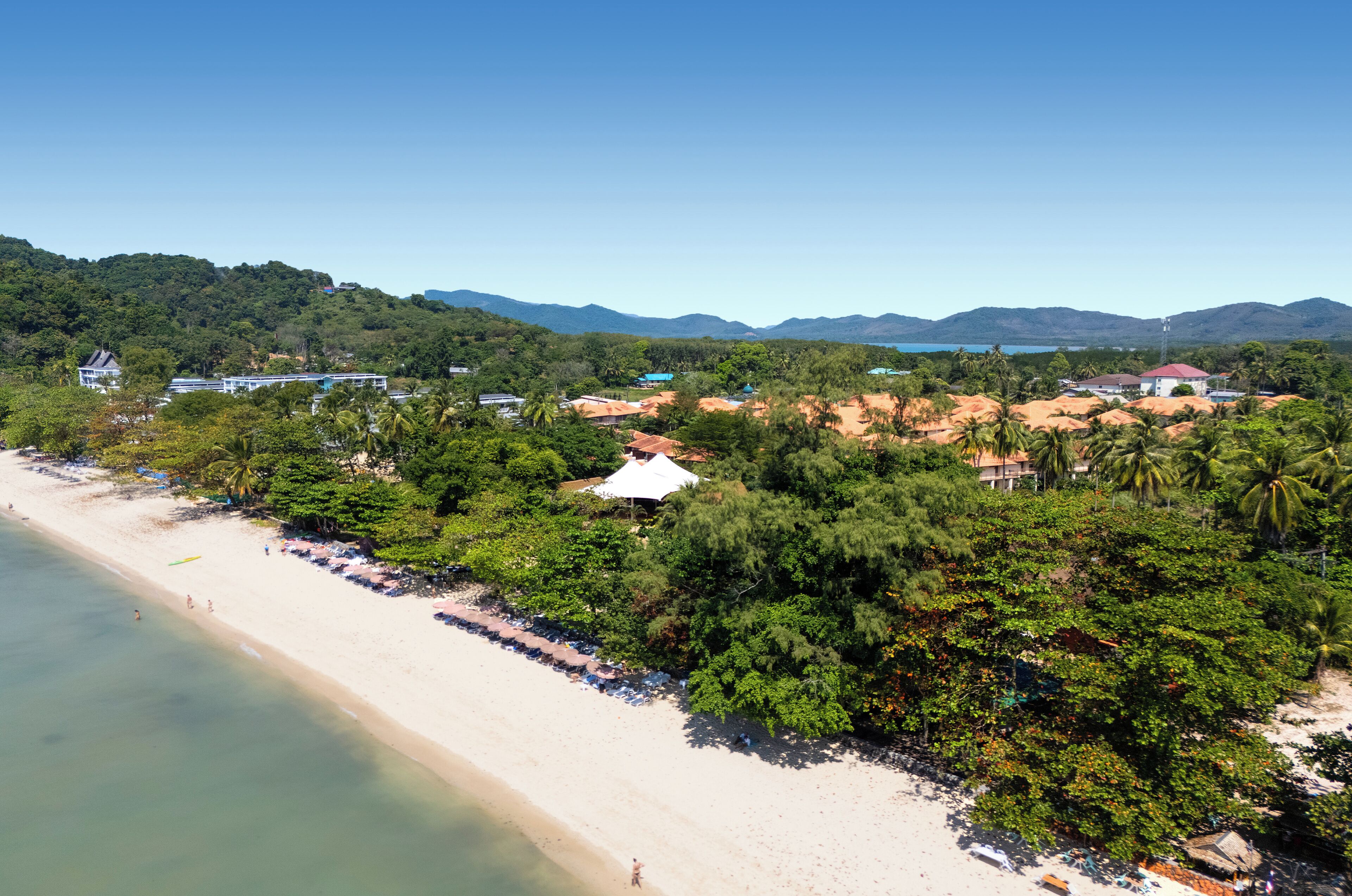 Un complejo turístico en la playa con una orilla arenosa, palmeras y montañas al fondo.
